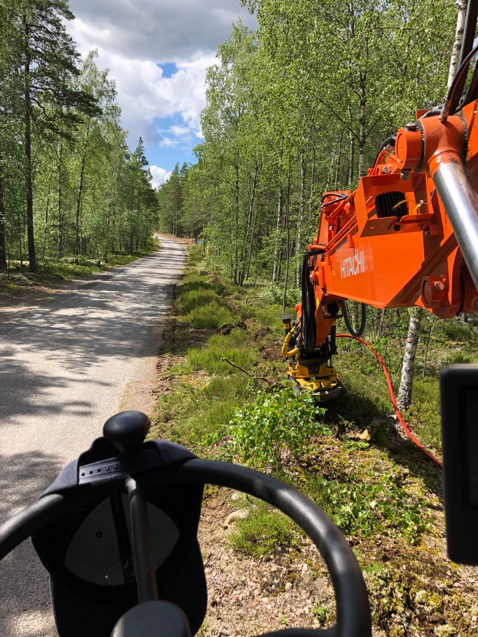 En grå grusväg kantas av skog och en orange grävmaskin.