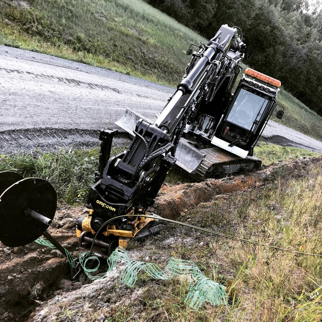 Grävmaskin arbetar i en grönskande gränsland med grön kabel och en stor spole.